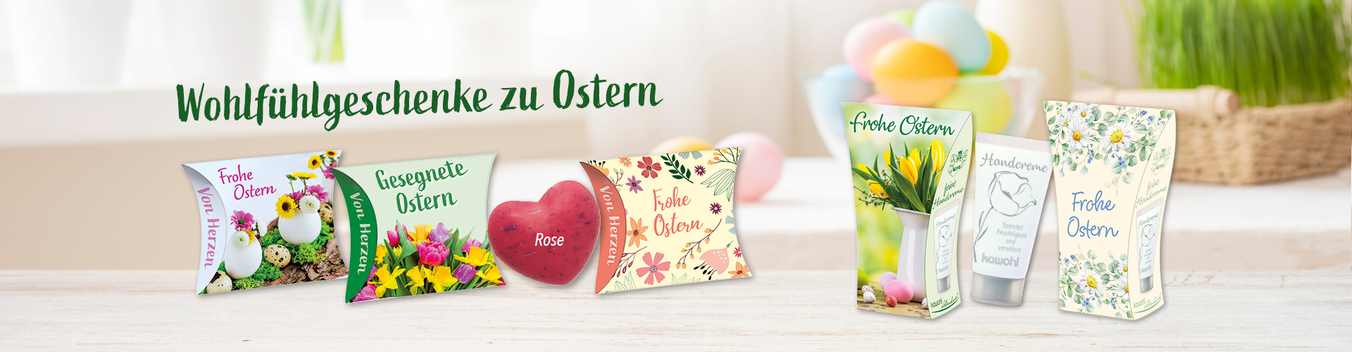 Foto von hellem Raum mit bunten Ostereiern und einer grünen Pflanze im Hintergrund. Verschiedene Handcreme- und Schafmilchseifen-Verpackungen mit Ostergrüßen sind abgebildet, über den Produkten steht der Text: „Wohlfühlgeschenke zu Ostern“.