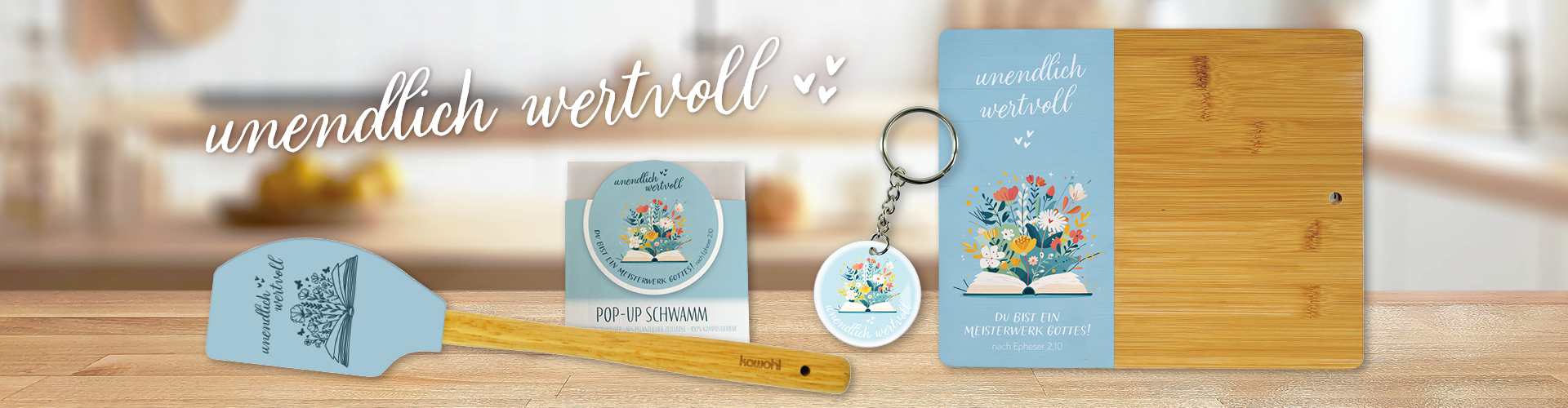 Produktkollektion "unendlich wertvoll" mit blauem Teigschaber, Pop-Up-Schwamm, Schlüsselanhänger und Bambus-Brett auf Holztisch.